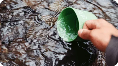 A man's hand holding a cup and dipping it into a clear shallow stream